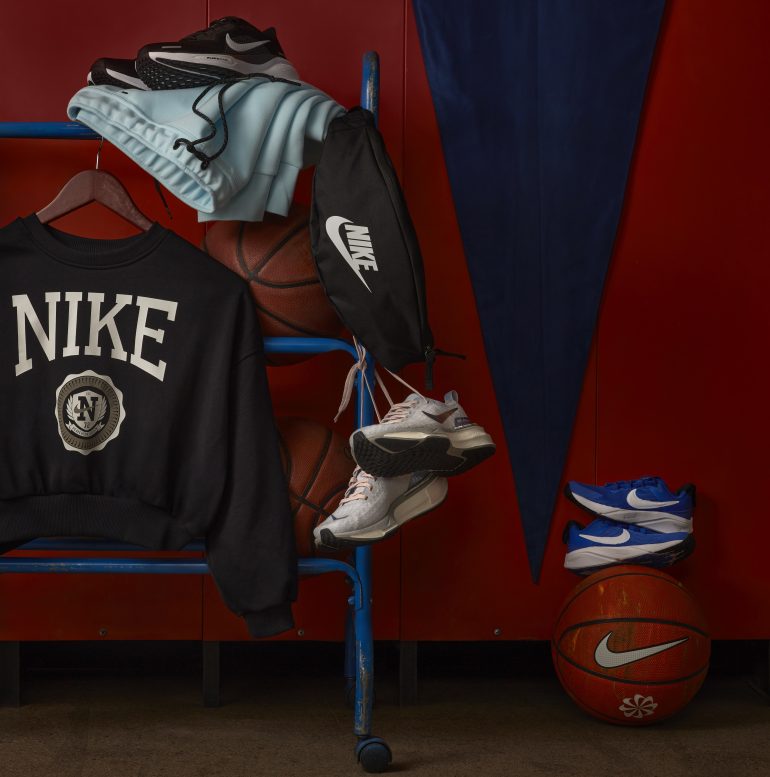 Nike gym class essentials hanging on a basketball rack with a pennant flag in the background, including a Nike crew sweatshirt, waist pack, sweatpants, three pairs of shoes and Nike basetballs