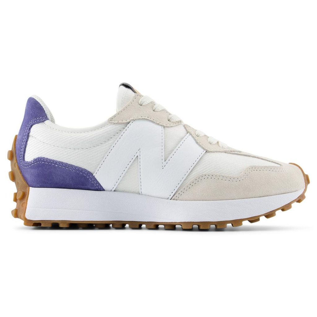Low-profile white/off-white New Balance sneakers with solid white foam sole and eggshell suede panel at the eyestay and toes, plus contrast blue suede patch at the heel. Brown gum rubber outsole that wraps up the toe and heel with small, circular lug pattern.