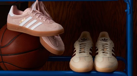 two pairs of adidas sneakers in muted pastel colours (light pink and tan/beige) sit on a blue metal ball rack with a classic brown basketball to the left and a dark wood wall in the background